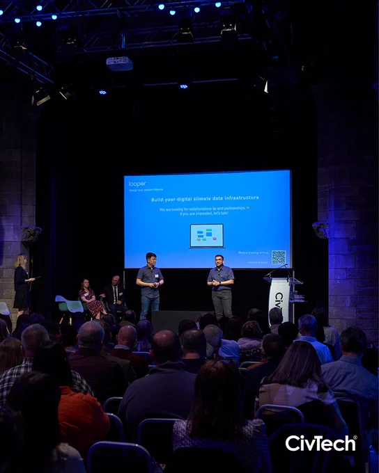 Two people stand on stage at the Assembly Roxy, presenting their solution at CivTech Demo Day in a dark room, facing rows of seated audience members.