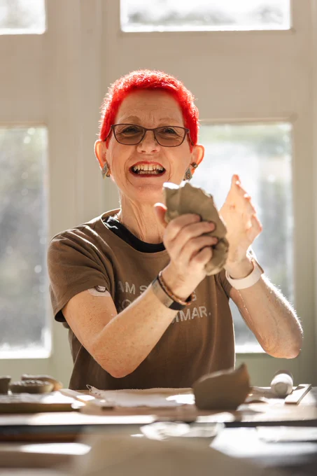 A woman with bright red hair and glasses smiles while shaping clay. She is in a pottery studio.