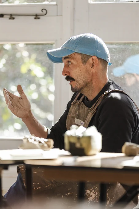 A man in a blue cap and apron gestures with his hand while sitting at a table with pottery supplies.
