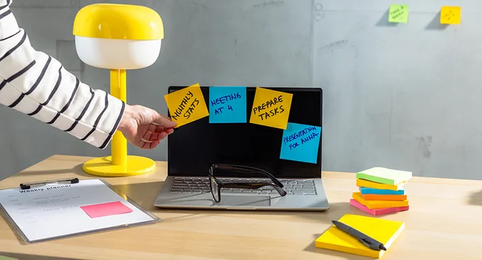 Person placing a yellow sticky note labeled 'Monthly Stats' on a laptop screen surrounded by other colorful notes and office supplies.