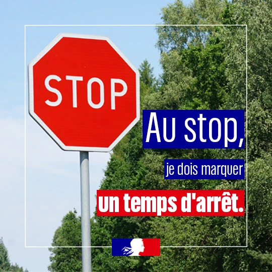 Un panneau STOP rouge au bord d’une route entourée d’arbres et de végétation. Un message de sécurité routière est affiché : « Au stop, je dois marquer un temps d’arrêt. » Le logo de la Marianne apparaît en bas de l’image.
