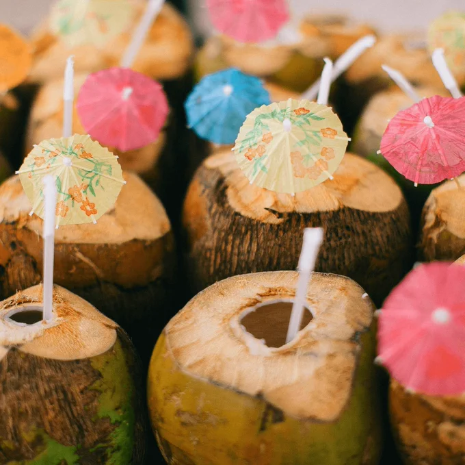 Coconuts holding cocktails with colorful umbrellas are lined up next to each other. Image by Lina Kivaka on Pexels.