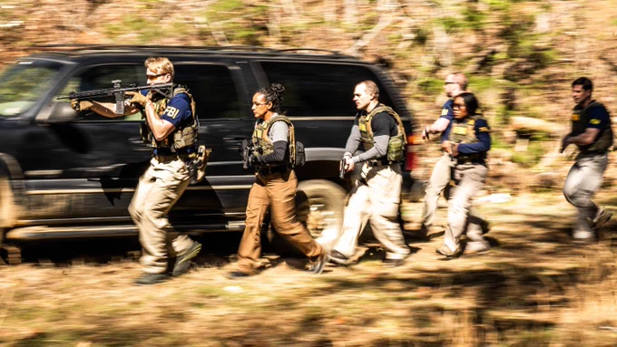 A team of FBI agents in tactical gear moving swiftly through a wooded area during a training exercise.
