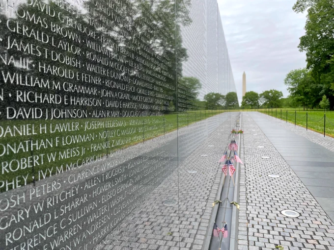 Close-up of the Vietnam Veterans Memorial wall with engraved names and small American flags placed along the pathway on a cloudy day.