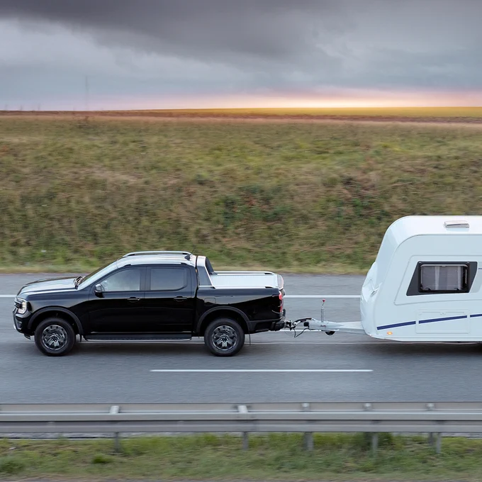 A black Ford Ranger towing a white caravan on a highway with grasslands and a cloudy sky in the background.