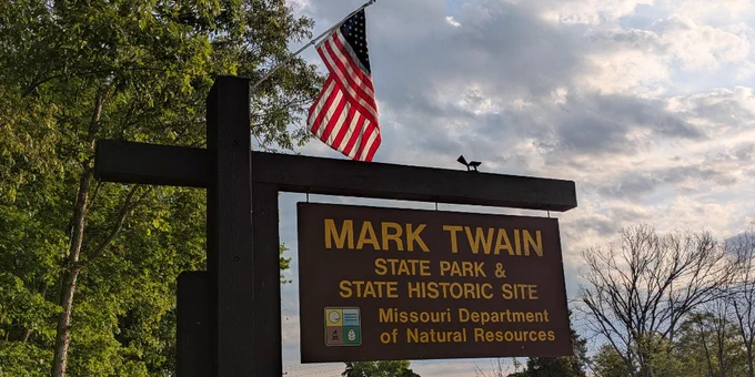 Wooden sign for Mark Twain State Park and Historic Site with an American flag above, surrounded by trees and a partly cloudy sky.