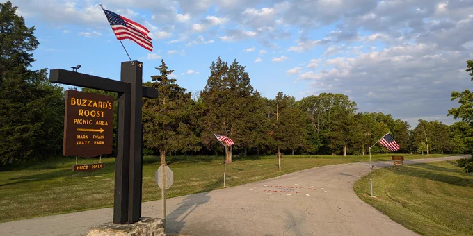 Entrance sign for Buzzard's Roost Picnic Area in a park with American flags lining the road under a partly cloudy sky.
