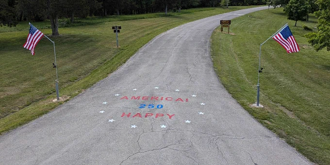 A curved road flanked by two American flags with a painted message on the pavement reading 'AMERICA! 250 HAPPY' surrounded by white stars.