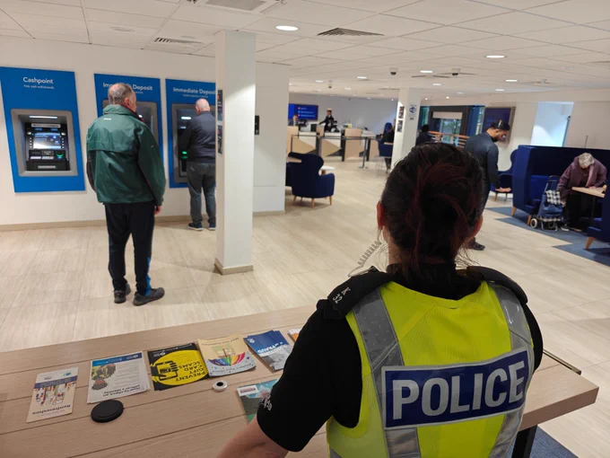 Police officer in uniform in foreground, within bank. ATMs in background.