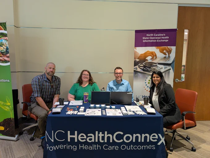 A group of four people is seated at a table covered with a dark blue cloth displaying the "NC HealthConnex" logo. The table is arranged with informational materials and promotional items. Behind them are two vertical banners, one mentioning "North Carolina's State-Operated Health Information Exchange." They're positioned in a modern indoor setting.