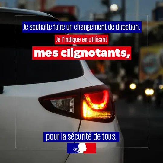 Un gros plan de l’arrière d’une voiture avec le clignotant droit allumé, dans une rue avec la nuit tombante. Un message de sécurité routière est affiché : « Je souhaite faire un changement de direction. Je l’indique en utilisant mes clignotants, pour la sécurité de tous. » Le logo de la Marianne est présent en bas.