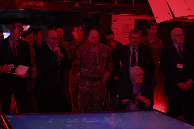 A group of people stand in a dimly lit room illuminated by red lighting. Some individuals are in military uniforms, while others are in suits. They are gathered around a display table, looking intently at its surface which shows a digital map or screen. The setting seems formal and focused.