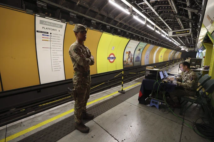 A person in military uniform stands on a London Underground platform next to a Jubilee Line sign. Another person sits at a table with electronic equipment. The platform is well-lit.
