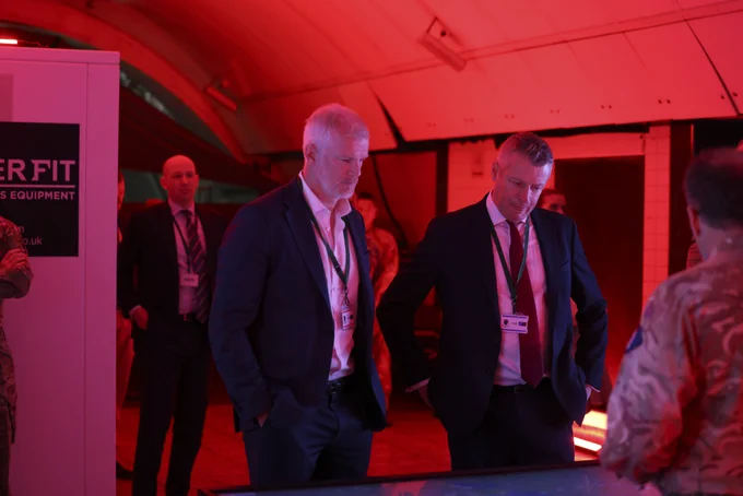 A group of people in business and military attire are gathered under red lighting. Two individuals in suits are attentively looking at a presentation screen. 