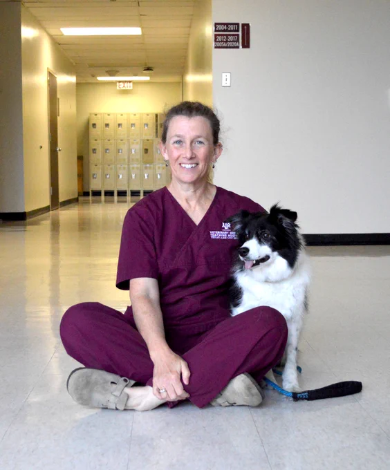 A person in scrubs sits cross-legged on a hallway floor next to a black and white dog.