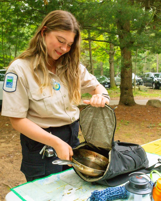 A park ranger is standing outdoors beside a table, opening a small bag containing pans. They are wearing a uniform with a patch that says "Ontario Parks." The setting is a wooded area with several parked cars in the background.