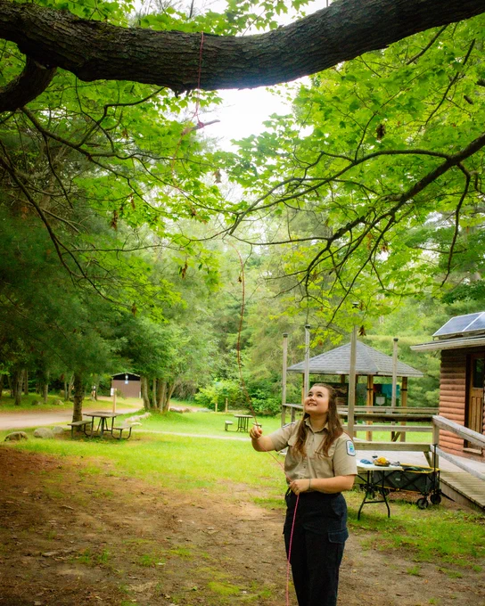 A person in a uniform is standing outdoors near a tree, holding a rope. The setting includes picnic tables, a wooden structure, and lush greenery.