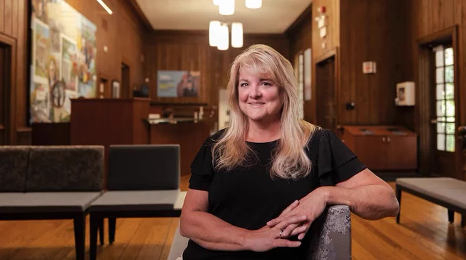 A person with long, light-colored hair is seated in a room with wooden walls. The room features framed pictures, a closed wooden door, and modern light fixtures on the ceiling.