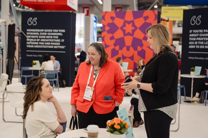 Three women chat around a table at a conference booth, with quote panels and a colorful geometric backdrop behind them.