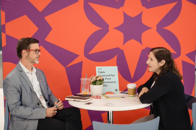 Two conference attendees talk at a small round table with coffee and notebooks against a bold orange-and-purple patterned wall.