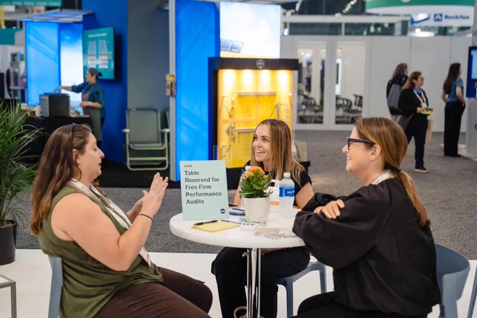 Three women chat at a round table in a trade show hall beside a sign reading, "Reserved for Free Firm Performance Audits."