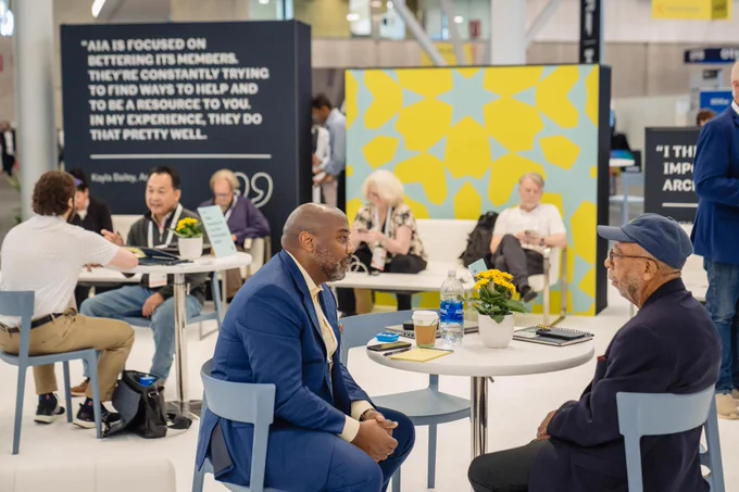 People chat at small tables in a bright conference lounge; two men converse in the foreground beside coffee, water, and flowers.