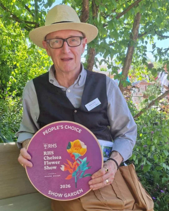 Our Head Gardener Barrie sitting in our Chelsea Garden underneath a tree holding the People's Choice Award. he is smiling 