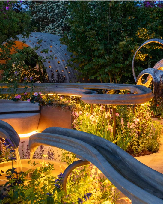 A photo from the centre of our garden at night time, the handrill is weaving through the plants and around the curvy pathway and the night shelter in the back is lit up, the lights are shining on the flowers bringing them to life