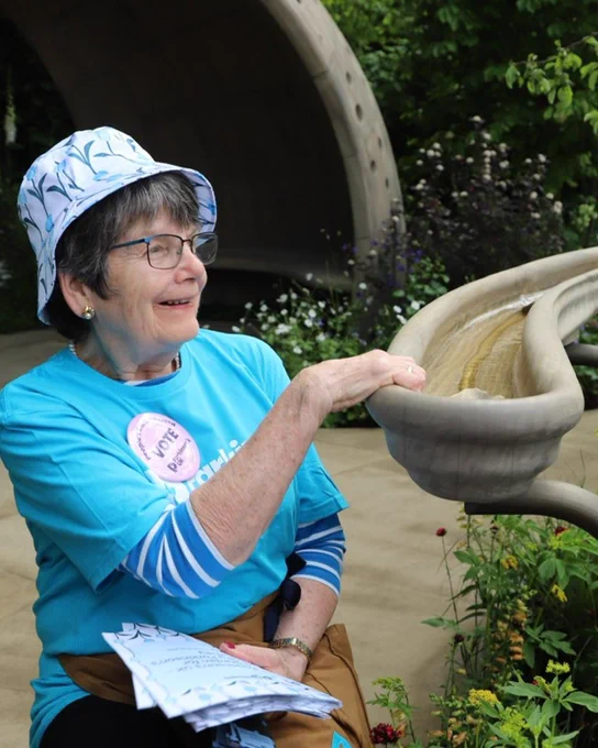 One of our head gardeners, Alison, is sitting on a chair in our garden, she is holding the handrill watching the water flow through. She is wearing a blue Parkinson's UK t-shirt, a tulip bucket hat and a vote for us badge