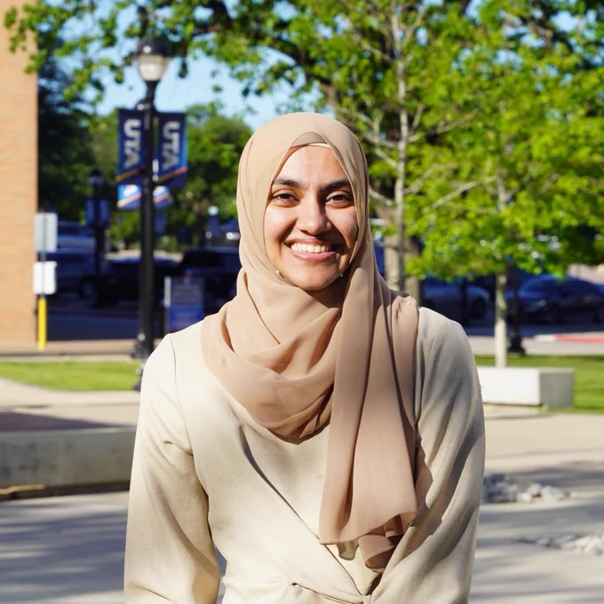 Photograph of University of Texas at Arlington Professor Tatheer Ajani wearing a tan-colored hijab and light-colored outfit standing outdoors on a sunny day with trees and campus buildings in the background.
