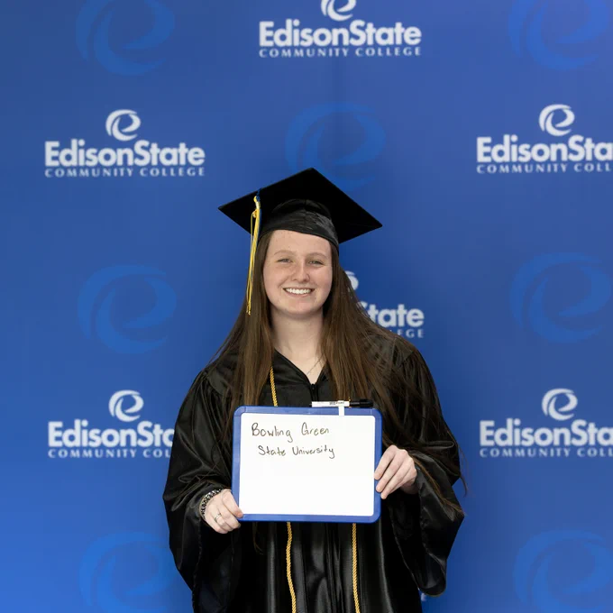 Graduate in black cap and gown holding a sign that reads Bowling Green State University against an Edison State Community College backdrop.