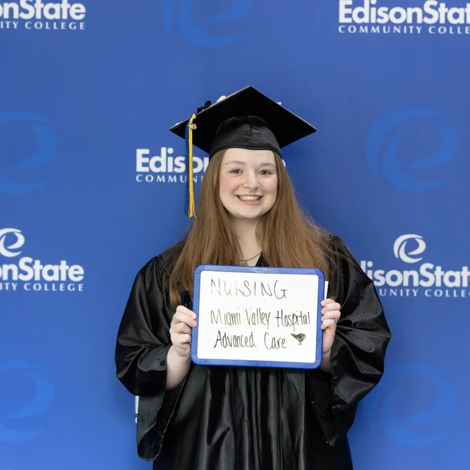 Graduate in cap and gown holding a sign announcing a nursing position at Miami Valley Hospital Advanced Care, standing before an Edison State Community College backdrop.