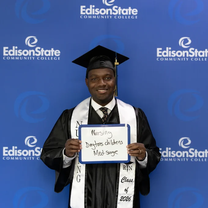 Graduate in cap and gown holding a sign listing Nursing: Dayton Children's, Med-Surge against Edison State Community College backdrop.