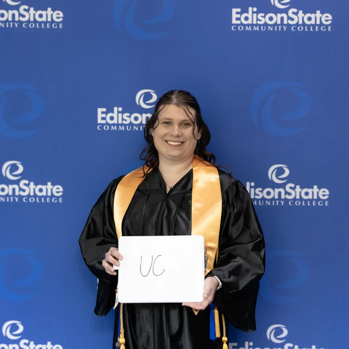Graduate in black cap and gown with gold stole holding a sign reading 'UC' in front of Edison State Community College backdrop.