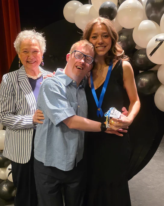 Three people are smiling in front of a backdrop with black and white balloons. The person in the middle is hugging the person holding a medal and an award.