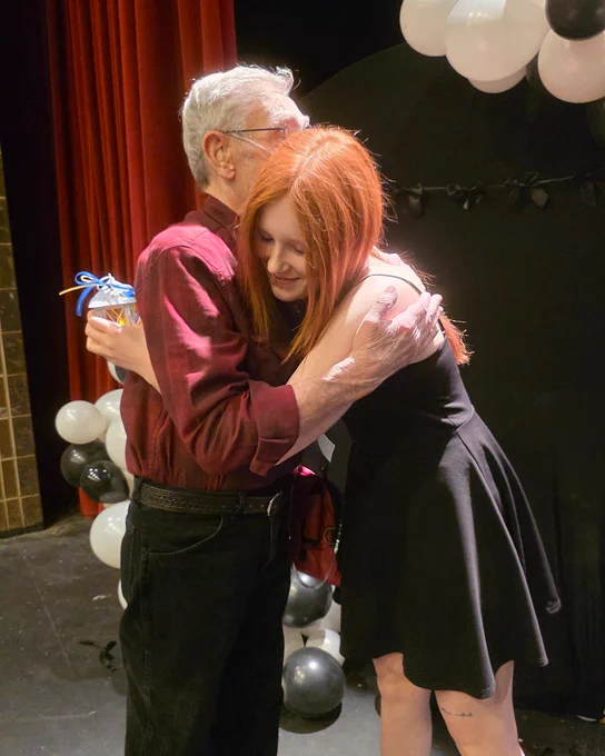 Two people embrace warmly at an event with a red curtain and black and white balloons in the background.