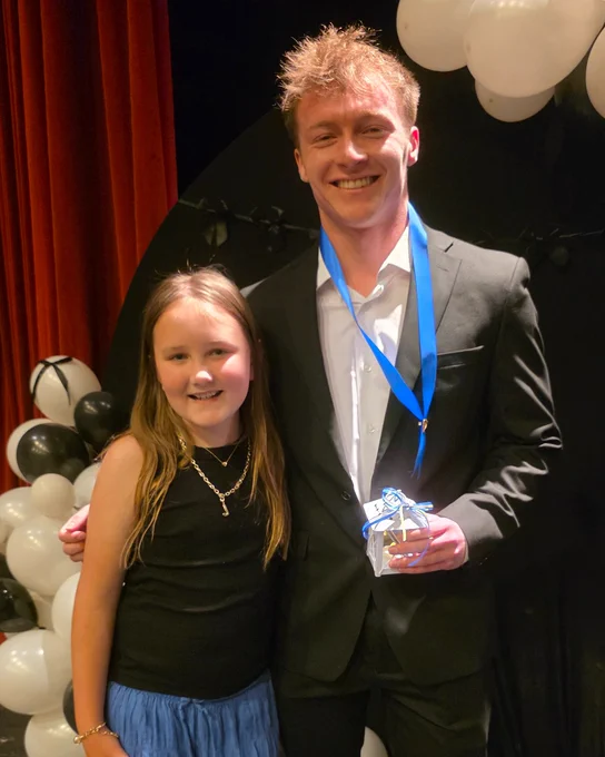 Two individuals are standing together, smiling. One is taller, wearing a suit with a blue ribbon and holding a small gift. The other is shorter, dressed in a black top and blue skirt. They are in front of red curtains and black-and-white balloons.