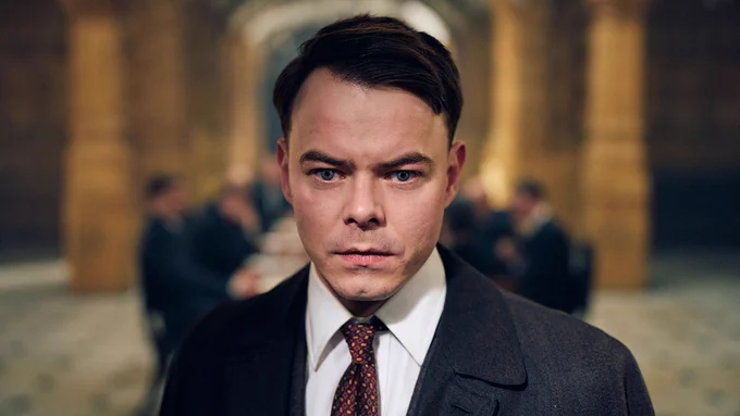 Charles Shelby (played by Charlie Heaton) is dressed in a dark overcoat, white shirt and patterned tie, looking directly ahead. Background shows a softly blurred, ornate interior hall with indistinct figures around a long table. “Coming to iPlayer” text is along the bottom.