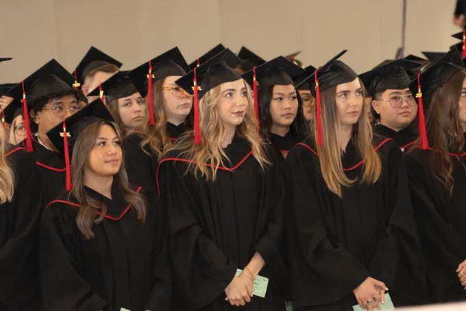 Alt Text: A large group of RRC Polytech graduates stand together in black graduation gowns and caps with red tassels during a convocation ceremony.