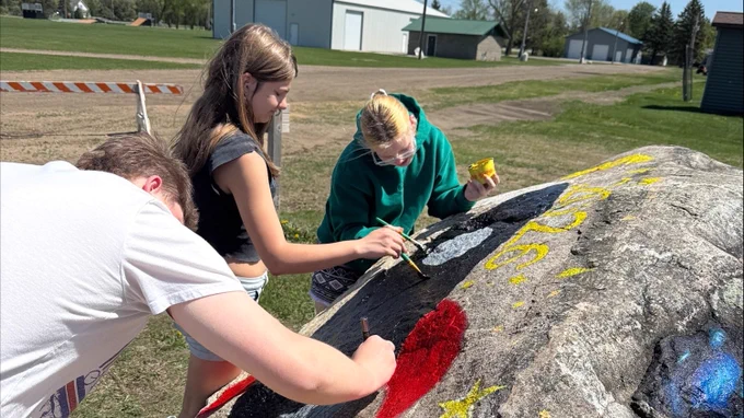 Three students paint a rocket on the big rock at the entrance of the ball field. 
