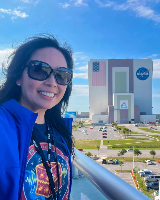 A woman wearing sunglasses, a blue jacket, a circular mission‑patch design t-shirt, and a NASA lanyard stands outdoors near a railing with a large NASA building in the background. The building features a massive painted American flag and the NASA logo. A parking lot, palm trees, and a bright sky with scattered clouds fill the scene.
