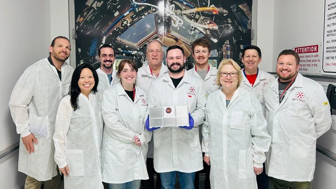 A group of 10 people in white lab coats stands together in a cleanroom setting. The person in the center holds a rectangular device with blue handles. Behind them is a large backdrop showing the interior of a spacecraft.