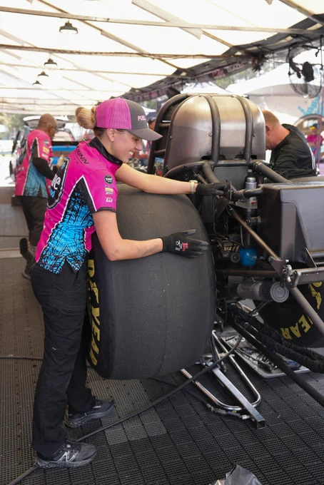 Smiling female racer in pink team shirt and cap holding a pink fuel product beside a blue-pink race car