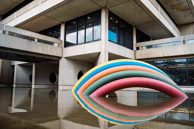 Colorful striped inflatable sculpture floating in a reflective pool under a modern concrete building.