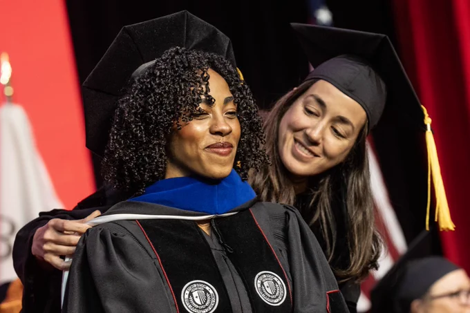 Two individuals in graduation attire are on stage. One person is placing an academic hood on the other. Both are wearing black caps and gowns with university emblems. An American flag and a red curtain are in the background.