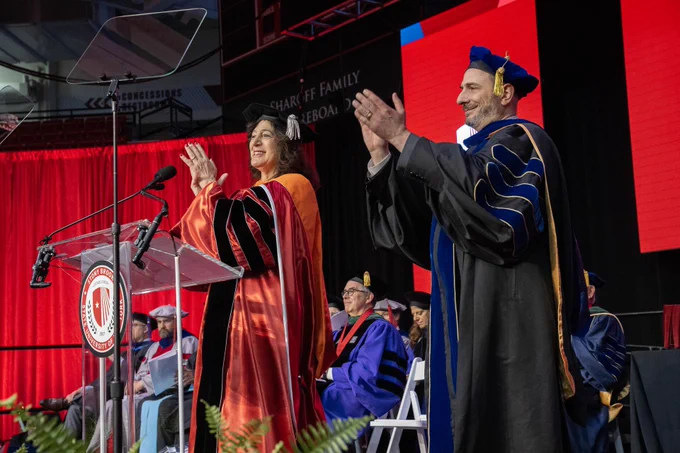 I am standing with Stony Brook University Provost Carl Lejuez. We are both clapping our hands in applause.  We are both in our academic regalia and standing on stage. A seal reading "Stony Brook University" is visible on the podium in front of us. Seated individuals in similar attire are in the background.