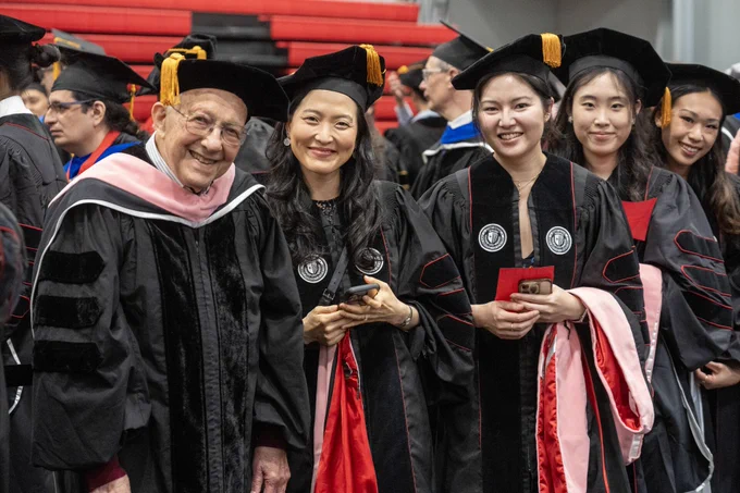 A group of people in academic regalia stand together, smiling at the Doctoral Hooding ceremony for a group photo. They are wearing black gowns with red and white details, as well as caps with tassels. In the background, more people in similar attire are visible.