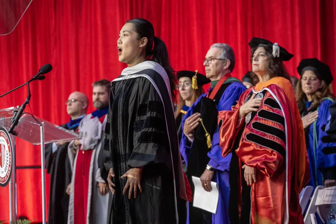 I am standing with Stony Brook University leadership on stage at the Doctoral Hooding ceremony. We are all dressed in in academic regalia. The backdrop is a vibrant red curtain. One person is singing at a microphone, while those on stage with me have our hands placed over our chests for the national anthem.