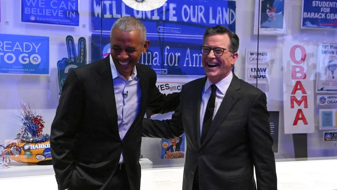 An image of Barack Obama and Stephen Colbert laughing in front of Obama campaign memorabilia at the Obama Presidential Library.