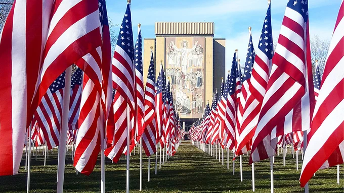 A view looking at the Word of Life mural at Hesburgh Library in the background, looking through many American flags planted in the Library lawn.